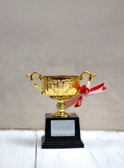 Little golden trophy with white ribbon and red ribbon on white wooden background. Copy space on trophy base.
