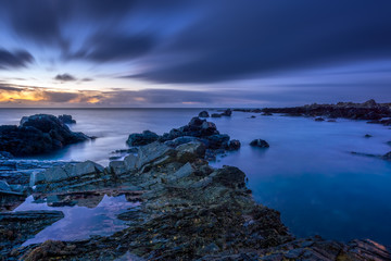 Twilight begins yielding to daylight at rocky coastline with blurred water and sky, long exposure photography. Northern Ireland