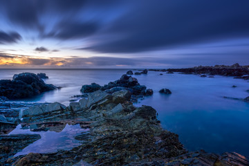 Twilight begins yielding to daylight at rocky coastline with blurred water and sky, long exposure...