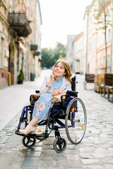 Fototapeta na wymiar attractive disabled blond young girl in blue dress sitting in a wheelchair, outdoors in the city
