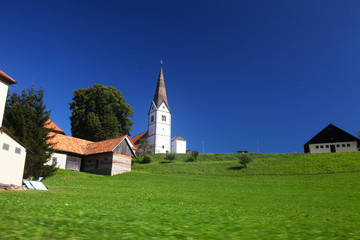 Fototapeta na wymiar Little white church in Slovenian countryside among lush green summer landscape and blue sky