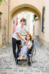 Young handsome smiling man and his pretty wife in wheelchair posing together on the background of old city center, arch of the ancient building. Love, disability concept