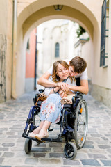 Couple in love in old city center. Pretty young girl with disease on a wheelchair and her lovely man, hugging her from the back