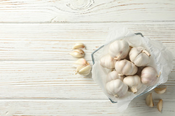 Glass bowl of fresh garlic bulbs, slices on white wooden background, top view. Space for text