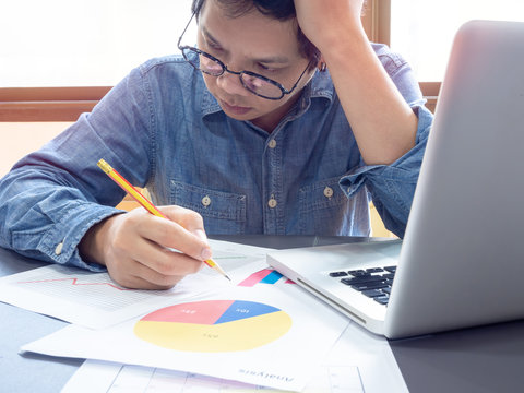 Asian businessman wearing blue shirt and glasses thinking with working with laptop computer on table near the window in office.