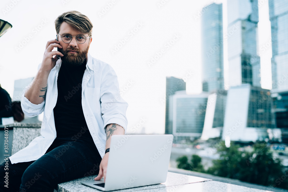Poster portrait of serious skilled male programmer in optical spectacles looking at camera while making imp