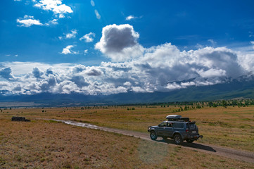 Fototapeta premium Steppe summer landscape against the background of distant mountains Adventure journey. Expedition car rides on a dirt road on the background of mountains, blue Sunny sky and storm front