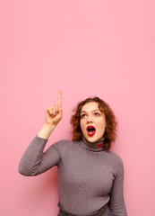 Shocked lady in casual clothes on curly red hair isolated on pink background, shows pointing finger up at copy space and looks up with surprised face, wearing gray sweater. Vertical photo.