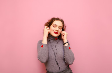 Attractive teen girl enjoys listening to music in headphones on a pink background with her eyes closed. Cute lady with bright makeup and red curly hair listens to music in headphones. Isolated.