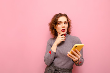 Pensive curly lady in gray clothes stands with smartphone in hand on pink background and looks away...
