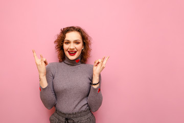 Happy cute girl with curly hair and casual clothes stands on a pink background and shows a heavy metal gesture, looks into the camera and smiles. Isolated. Rock music concept.
