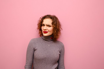 Cute girl with curly hair and casual clothes looks suspiciously into the camera on a pink...