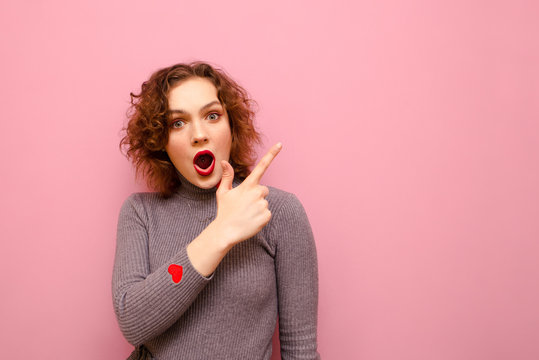 Shocked young lady points her finger at copy space and looks into camera with emotional face. Surprised curly lady stands on a pink background and points her finger at an empty place