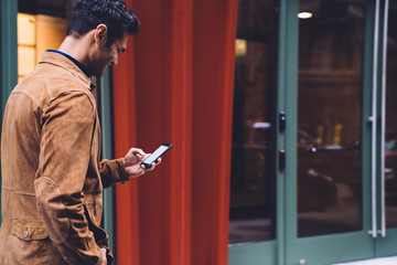 Young smiling male in stylish clothes walking while using smartphone
