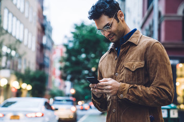 Captivated ethnic young man in jacket texting on street