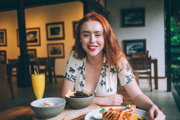 Cheerful redhead woman sitting at cafe table with various food