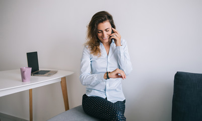 Smiling young woman checking time on wristwatch while talking on phone
