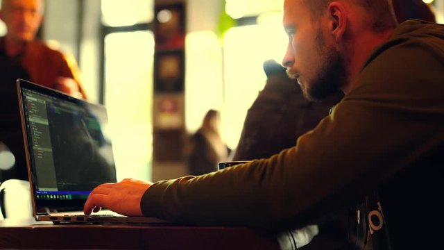 side view man programmer freelancer sitting in cafe working using laptop, man types, codes, Writes Emails, Surfs the Internet, Designs Software. cup of coffee on table