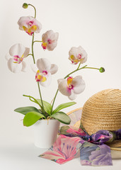 Bodegón sobre fondo blanco de un sombrero de paja con lazo en tonos violetas y maceta con orquídeas blancas artificiales 