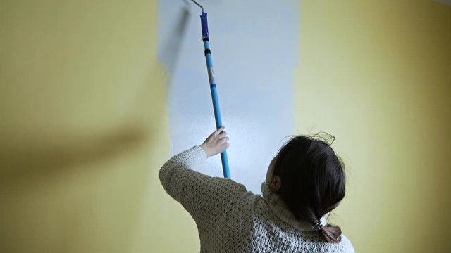 Joyful self-taught girl with a construction roller paints a yellow wall with gray paint. Independent apartment repair. Fresh repairs after moving to a new apartment