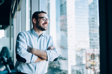 Cheerful male entrepreneur with crossed hands standing near office window view and smiling during...