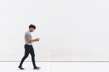 Handsome young curly man in casual outfit browsing smartphone while walking on marble passage