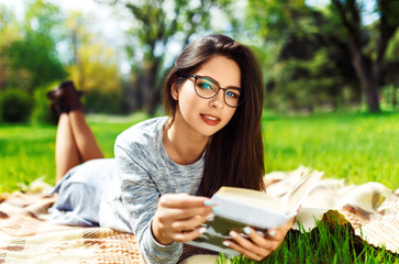 Beautiful  girl in eyeglasses reading book in the summer park. Young student girl  lies on green...