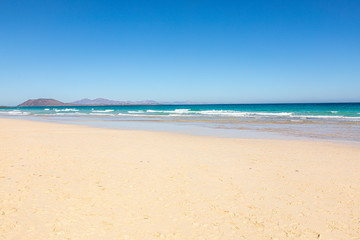 Fototapeta na wymiar Beach on Fuerteventura canary islands with the beach in front and the sea with blue azure aquamarine turquoise colors and a vulcano in the background blue sky