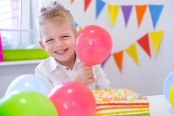 Fototapeta na wymiar Portrait of blonde caucasian boy smiling at camera near birthday rainbow cake. Festive colorful background with balloons.
