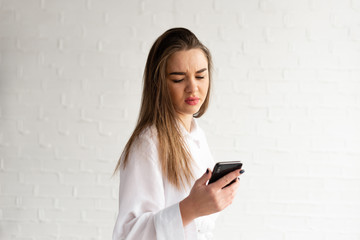 A young woman in a white blouse looks at her phone, emotion of surprise and misunderstanding
