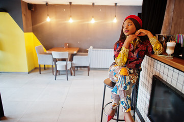 Enthusiastic african american woman in trendy coloured outfit with red beret chilling in cozy cafe, sitting on bar counter.