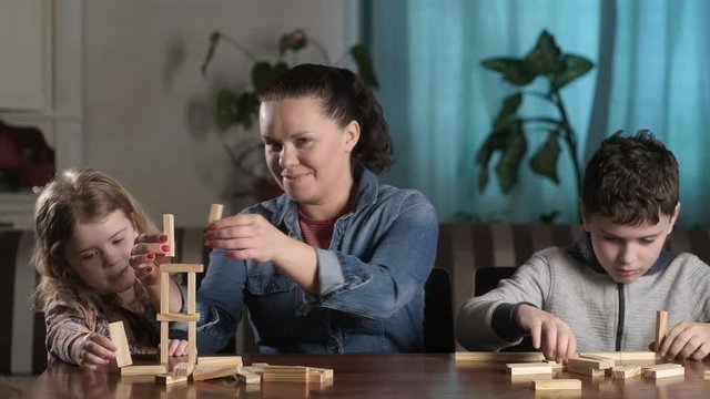 beautiful young mother helps her son and daughter build  tower of wooden blocks. Children are focused on game. family have fun playing with wooden blocks. Educational Board games for the whole family.