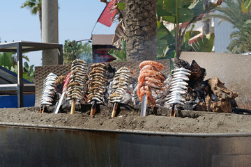 Close-up of sardines and scampi grilled with wood on spits on a boat shaped silver grill at the...