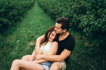 Woman with long hair and man sitting between currant bushes, hugging and kissing. Background of green currant plantations