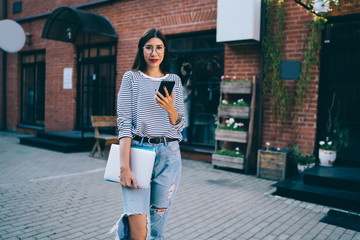 Half length portrait of successful female graphic designer holding laptop computer for distance job and modern cellular device for online browsing and looking at camera during free time at urbanity