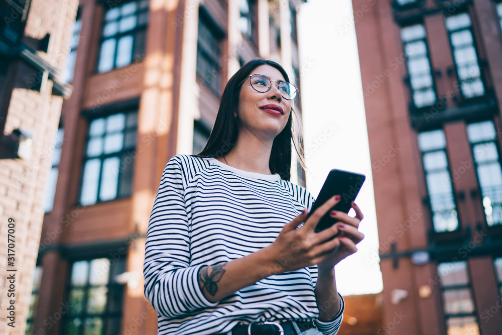 Aufkleber below view of millennial blogger with cellphone in hands looking away and smiling enjoying time for 