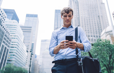 Below view of employer reading sms on modern mobile phone while standing at urban setting in downtown and using 4g wireless internet connection for communicate, intelligent businessman chatting