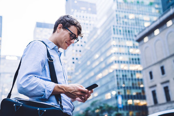 Young employee in glasses texting on phone smiling standing at street