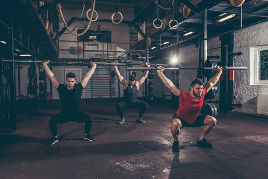 Group of three young friends fit muscular fitness guys having fun by hard core cross workout training in the home made gym with barbells and squats selective focus with film grain