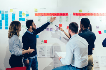 Young multiethnic business partners having discussion while analyzing wall with sticky notes at...