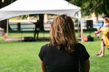 Fototapeta premium Closeup rear view of woman with blonde hair wearing black t-shirt on grassy lawn against tent. in park at picnic on a bright sunny day