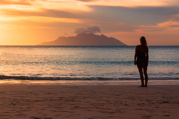 Young girl standing on the beach looking at the island far away during sunset , girl silhouette from behind , orange and blue sunset on Seychelles Beau vallon beach