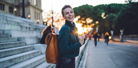 Half length portrait of cheerful hipster girl with cellphone device in hands smiling at camera during online communication with followers from web blog, happy tourist with travel backpack rejoicing