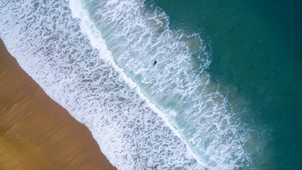 Top view of a person surfing in a long sand beach. In the image we can see the ocean with big waves. 