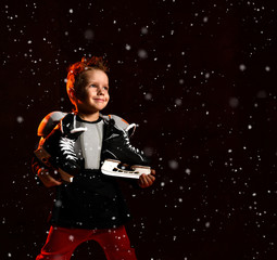Blond boy in hockey uniform with hockey skates on neck standing and looking at camera over dark background