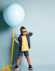 Blond boy in stylish casual clothing, sneakers and sunglasses standing near blue air balloon and looking at camera