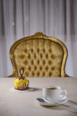 Mini caramel cream cake white cup of coffee, spoon,  on a marble table in the background antique gold chair. Focus on the mini cake