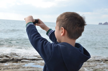 boy schoolboy or teenager in a dark blue tracksuit walks along the sea, takes a selfie or shoots the sea on an action camera, on the sea waves, stone coast, wind