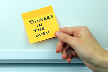 A hand putting a sticky on on the fridge door with the message dinners in the oven.