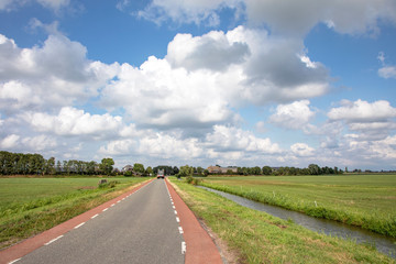 Fototapeta na wymiar Road in Holland with red cycle path on both sides, perspective, under heavy cloudy skies and between green meadows, agricultural vehicle drives at the end of the road